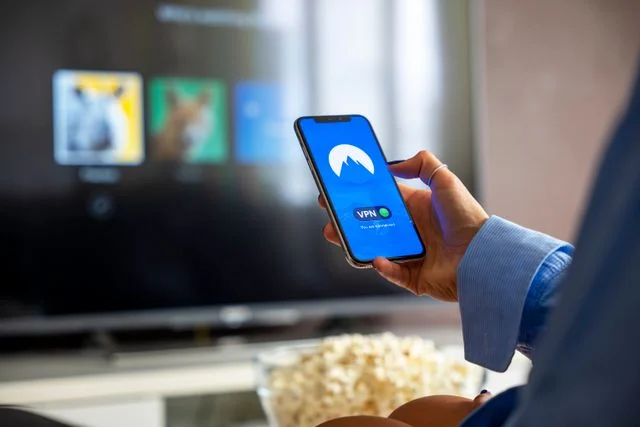 NordVPN review 2026: testing the app for streaming, gaming and AI tools on phone and TV Hand holding a smartphone with the NordVPN app connected while a television streams in the background